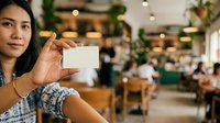 Woman holding blank white business card in modern cafe with plants and warm lighting, ideal for branding and corporate marketing materials.