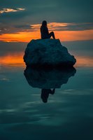 Woman with long dark hair sitting on rocky boulder in calm ocean water at sunset, silhouetted against golden and orange sky with dramatic clouds and reflection.