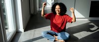 Young African American woman celebrating success with raised arms while sitting cross-legged on floor with laptop and documents in bright modern office space.