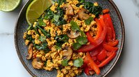 Fresh tofu scramble with sauteed mushrooms, kale, red bell peppers and lime wedges served in gray ceramic bowl on marble surface, healthy vegan breakfast.