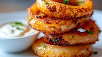 Crispy golden onion rings stacked in layers with fresh herbs and creamy dipping sauce, close-up view showing texture and caramelization on white plate.