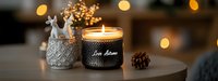 Glowing autumn scented candle in black glass jar with decorative white ceramic holder and pine cone, creating cozy atmosphere against blurred bokeh lights.