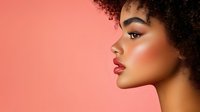 Side profile of young African American woman with natural curly afro hair, red lipstick, and glowing makeup against coral pink background. Fashion beauty portrait.