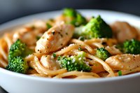 Creamy chicken pasta with fresh broccoli florets and black pepper in white ceramic bowl, selective focus on tender meat pieces and vibrant green vegetables.
