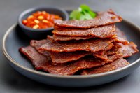 Crispy beef jerky strips served on dark plate with spicy dipping sauce and fresh herbs for snacking and appetizer presentations.
