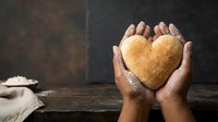 Hands gently holding golden heart-shaped bread on rustic wooden table. Warm bakery concept for love, care, and homemade cooking themes.