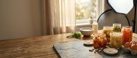 Homemade preserves and pickled vegetables in glass jars on rustic wooden kitchen table with natural sunlight streaming through window curtains.