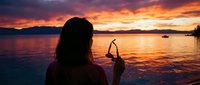Woman with long dark hair photographed from behind at sunset, holding smartphone to capture golden hour reflection on calm water with boats and mountains in distance.