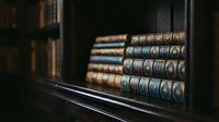Antique leather-bound books with ornate spines arranged on dark wooden shelf in library or study room, showcasing classic literature collection and vintage bookcase interior design.
