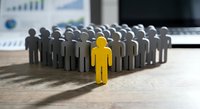 Yellow figure standing out from crowd of gray people on office desk with business charts and laptop in background representing leadership and uniqueness.