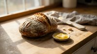 Artisan sourdough loaf with crispy crust and scoring marks beside melted butter on wooden surface, ideal for bakery, food, and culinary content.