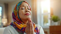 African woman wearing colorful hijab praying at home with peaceful expression, eyes closed and hands clasped in spiritual meditation moment.
