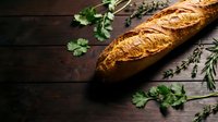 Freshly baked artisan bread loaf with golden crust on rustic wooden table surrounded by fresh herbs including parsley and rosemary for culinary cooking.