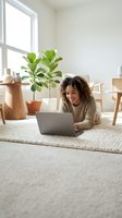 Young woman with curly dark hair and medium skin tone working on laptop in bright, minimalist home office with potted plant, wooden furniture, and natural light from window.