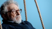 Thoughtful elderly Caucasian man with gray beard and glasses sitting on rope swing against blue sky background, contemplating life in peaceful moment.