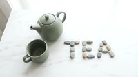 Sage green ceramic teapot and cup on marble surface beside the word TEA spelled with natural pebble stones, minimalist style.