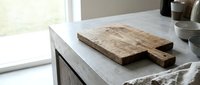 Rustic wooden cutting board with handle on white marble kitchen counter near window with natural light for culinary food preparation and cooking.