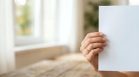Person holding blank white card or paper indoors with natural window light, suitable for mockups, advertising, and promotional designs.