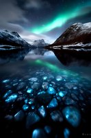 Northern lights dance over snow-capped mountains, reflecting in crystal-clear lake water with glowing stones beneath surface creating magical winter landscape at night.