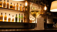 Glass of white wine on wooden bar counter with blurred bottles and warm lighting creating cozy restaurant atmosphere for dining and hospitality concepts.