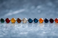 Colorful gemstone rings lined up in row on textured surface against blurred blue background, featuring emerald, ruby, topaz, and other precious stones in silver settings.
