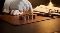 Business professional counting coins on wooden desk with documents and glasses in warm office lighting for financial planning and budgeting concepts.
