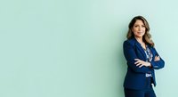 Young woman with wavy brown hair and olive complexion wearing navy blazer with arms crossed against mint green background, suitable for corporate, business, and professional portraits.