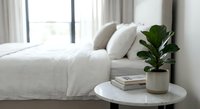 Minimalist bedroom interior with white bedding, fiddle leaf fig plant in ceramic pot on round side table near window with natural light streaming through curtains.