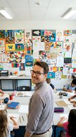 Male elementary school teacher with glasses smiling in colorful classroom with student artwork displayed on walls during art lesson with children.