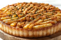 Caramelized pear tart with flaky pastry crust and golden brown fruit arranged in concentric circles, garnished with fresh herbs on wooden serving board.
