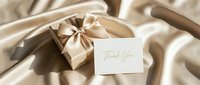 Elegant gold gift box with satin ribbon and thank you card on luxurious beige silk fabric, perfect for holiday, wedding, or appreciation themes.
