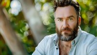 Confident mature bearded man with intense gaze in casual denim shirt against blurred natural bokeh background, expressing wisdom and masculinity.