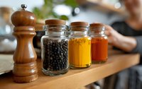 Wooden pepper mill and glass jars with black peppercorns, turmeric, and paprika spices on kitchen shelf, cooking ingredients for culinary use.
