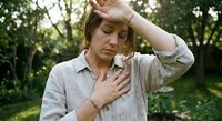 Middle-aged Caucasian woman experiencing chest pain and dizziness outdoors, holding head and chest suggesting heart attack or cardiovascular distress symptoms.