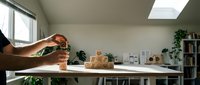 Hands holding small potted plant over modern white desk in bright home office workspace with natural lighting and green plants creating productive environment.