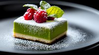 Green matcha cheesecake square topped with fresh raspberries and mint leaves, dusted with powdered sugar on dark plate, close up view of gourmet dessert.