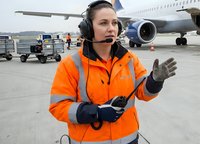 Female airport ground crew worker in orange safety vest with headset directing aircraft operations on tarmac with commercial airplane in background.