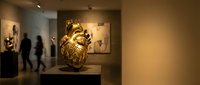 Golden ornate vase displayed on pedestal in modern art gallery with visitors viewing contemporary artwork exhibitions in warm ambient lighting.