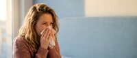 Woman with long wavy brown hair and fair complexion blowing nose into tissue while sitting indoors, depicting cold, flu, or allergy symptoms during daytime.