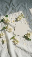 Blank white card surrounded by fresh white jasmine flowers and green leaves on crumpled linen fabric with natural sunlight shadows, perfect for wedding invitations, greeting cards, and stationery design.