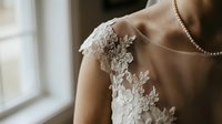 Elegant bride in white lace wedding dress with floral embroidery and pearl necklace by window. Bridal fashion and wedding day preparation concept.