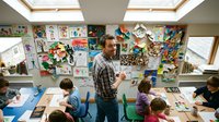 Male teacher with beard standing in bright elementary classroom with skylights, surrounded by children at desks with colorful artwork displayed on walls.