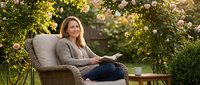 Relaxed middle-aged Caucasian woman reading book in wicker chair surrounded by blooming rose garden during golden hour sunset for lifestyle content.