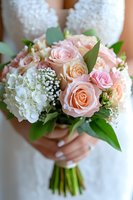 Bride holding elegant wedding bouquet with blush pink roses, white hydrangeas, baby's breath and green foliage against white lace dress background.
