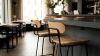 Modern wooden chairs with curved backs and metal legs positioned at a concrete bar counter in a bright, minimalist cafe or restaurant interior with natural light and contemporary decor.