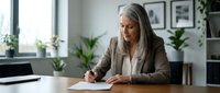 Professional mature businesswoman with gray hair writing documents at modern office desk with plants and natural lighting for corporate presentations.