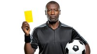 African American male referee in black uniform holding yellow card and soccer ball, showing serious expression against white background for sports design.