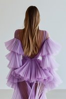 Young woman with long brown hair wearing lavender pleated tulle gown with dramatic ruffled sleeves, photographed from behind against white background.