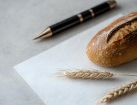 Fresh crusty bread loaf with golden wheat ears and elegant pen on white textured background, minimalist food composition for bakery marketing and design.