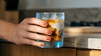 Woman holding glass of whiskey with ice on wooden table, close-up of hand and drink for beverage, lifestyle, and hospitality concepts.